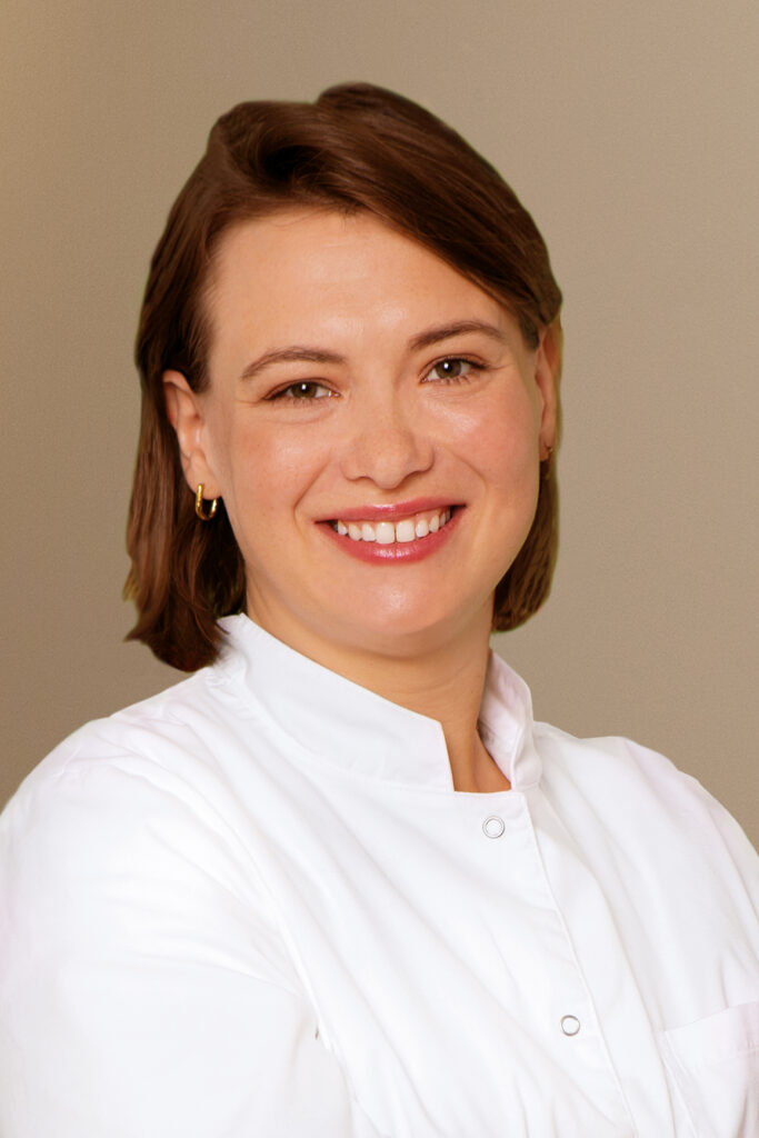 Eine lächelnde Frau mit braunen Haaren trägt einen weißen Arztkittel vor hellem Hintergrund