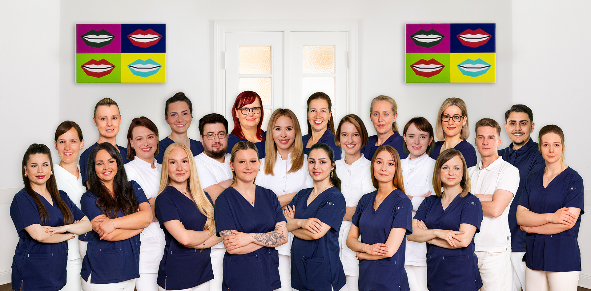 Eine Gruppe von zwanzig Menschen in weißer und blauer Kleidung lächelt freundlich vor bunter Wandkunst