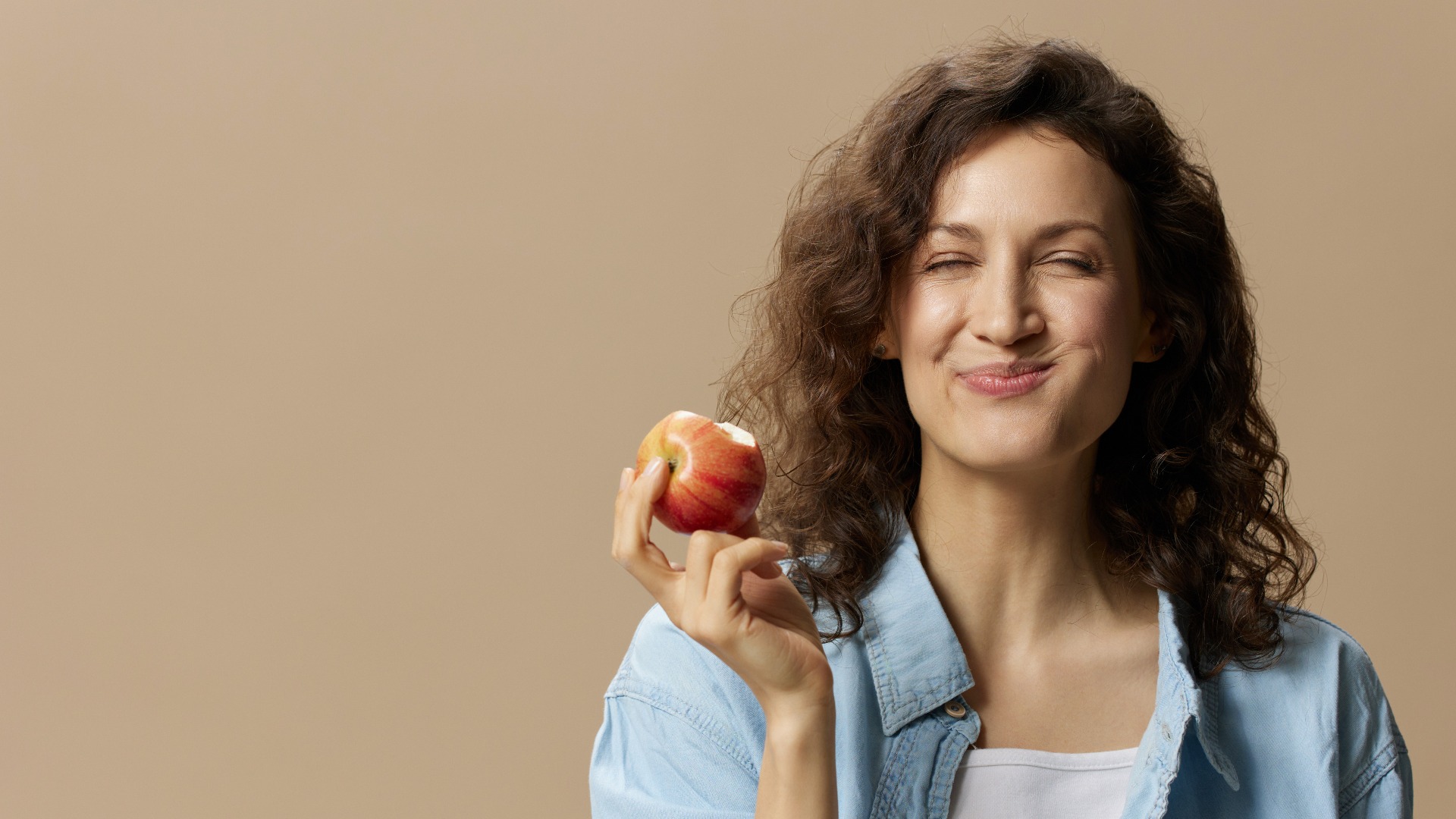 Lockige Frau knabbert an einem Apfel vor beigem Pastellhintergrund.